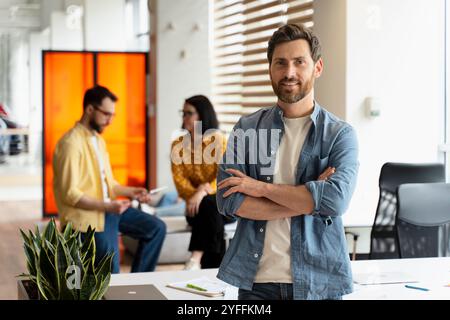 Smiling businessman posing with arms crossed in modern office while his colleagues are working in background Stock Photo