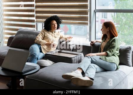 Two young female colleagues are enjoying their break time in the office, casually chatting and sharing a moment of relaxation on a comfortable sofa Stock Photo