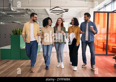 Group of five happy coworkers are having a lively discussion while walking together through their modern office space Stock Photo