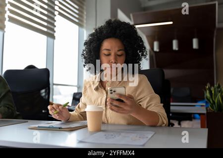 Focused black businesswoman is using her phone and taking notes at her desk in a modern office Stock Photo