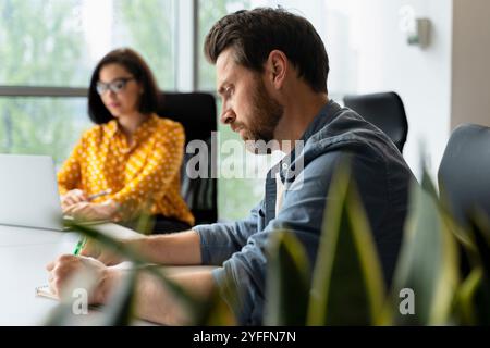 Focused manager taking notes during collaborative meeting with female colleague in modern office, working on planning and strategizing with laptop and Stock Photo