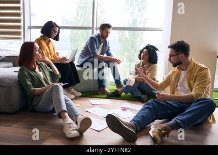 Young man leading brainstorming session with diverse group of colleagues sitting on the floor, discussing new ideas and developing strategy for projec Stock Photo