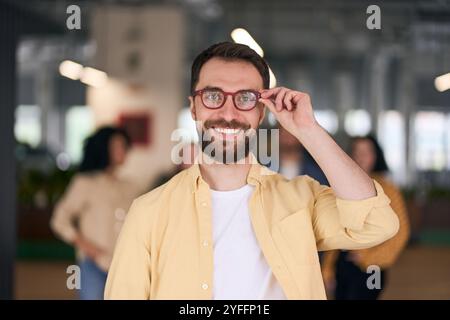 Bearded professional man wearing eyeglasses smiling confidently in a modern office with colleagues working in background Stock Photo