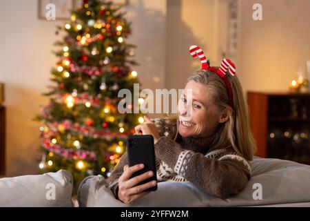 Smiling senior woman using smartphone at home, Christmas tree glowing behind Stock Photo