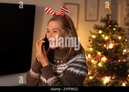Smiling senior woman wearing festive headband talking on phone near Christmas tree, at home Stock Photo
