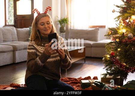 Smiling senior woman wearing festive headband using smartphone near Christmas tree at home Stock Photo