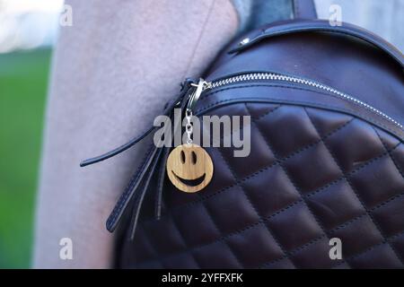Woman with wooden keychain in shape of smiley face and black leather decor on her backpack outdoors, closeup. Selective focus Stock Photo