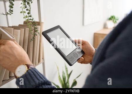 Young man with e-reader and books on shelf at home, closeup Stock Photo
