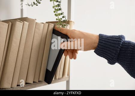 Young man taking e-reader from shelf with books at home, closeup Stock Photo