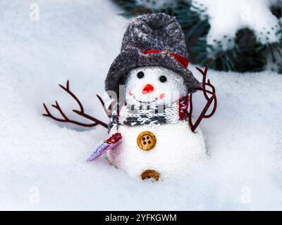 Small snowman figurine sitting in deep fresh snow with evergreen branches in the background. Stock Photo