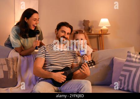 Happy family with microphones and ukulele singing karaoke at home in evening Stock Photo