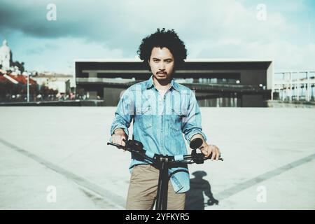 Young man with curly hair in a denim shirt standing outdoors, holding the handlebars of a modern electric scooter. Contemporary architecture and cloud Stock Photo