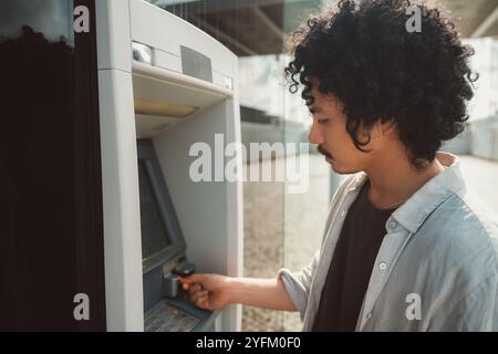 A wide-angle shot of a young man with curly hair and a mustache stands at an outdoor ATM, focused on the screen as he inserts his bank card. Dressed c Stock Photo