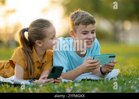 Positive children boy and girl playing online games on their mobile phones with friends outdoors in summer park Stock Photo