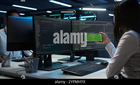 Zoom in on green screen mobile phone used by data center employee troubleshooting issues. Engineer uses mockup smartphone device in server hub, establishing performance benchmarks, camera B Stock Photo