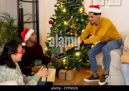 multiracial friends exchanging gifts by Christmas tree, wearing festive hats and smiling, at home Stock Photo