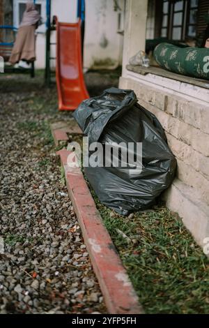 A black garbage bag placed against a stone wall, on a ground covered with small pebbles and patches of grass. An orange slide is visible in the backgr Stock Photo