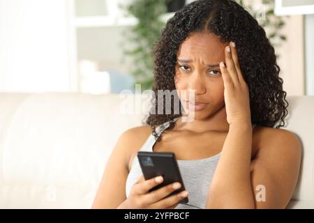 Worried black woman holding phone looking at camera at home Stock Photo