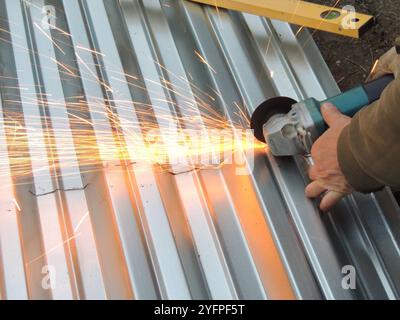 hands with grinder sawing corrugated grey shiny sheet of iron with bright sparks, grinding metal covering material on the ground using hand tool Stock Photo