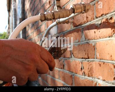 hand brings gas wrench to system of white thin plastic pipes coming out of red brick wall outdoor, repair with hand tool of pipeline on street Stock Photo