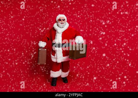 Photo of funny excited santa claus wear red headwear hat dark glasses smiling hurrying check in isolated red color background Stock Photo