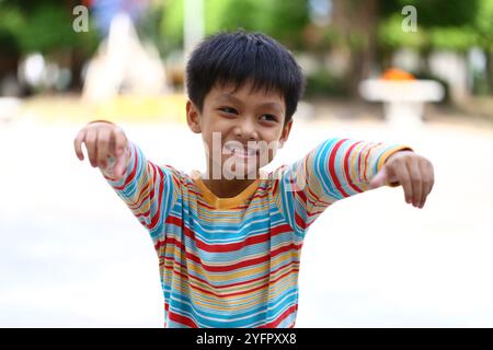 A cheerful boy wearing a colorful striped shirt enjoys a sunny day outdoors. His joyful expression and playful gesture capture the essence of childhoo Stock Photo