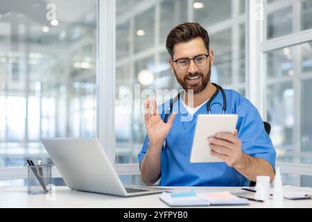 Doctor in blue scrubs using tablet for online consultation, smiling and engaged. Modern office setting with laptop and paperwork on desk, showcasing telemedicine and digital healthcare. Stock Photo