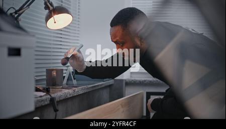 Murder or robbery case: African American policeman finds fingerprints on cup belonging to crime victim using brush and magnetic powder. Male forensic scientist works with evidence at crime scene. Stock Photo