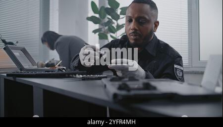 Murder case: Close up of African American police officer finding fingerprints on knife using brush and magnetic powder. Female criminalist works in the background. Investigation team at crime scene. Stock Photo