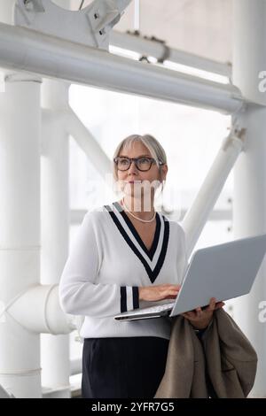 An older woman in a white sweater works on her laptop while standing indoors in a modern space with industrial features Stock Photo