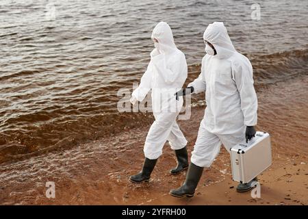 High angle view of two scientists wearing protective suits while walking along water and carrying toolkit case, copy space Stock Photo