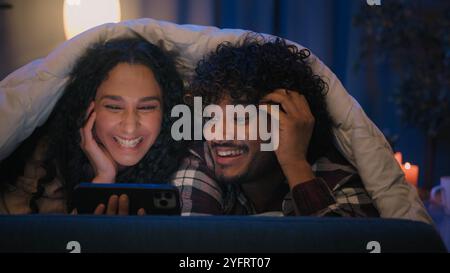 Multiracial couple man and woman happy cover with blanket under duvet at night evening home using mobile phone online browsing talking discuss video Stock Photo