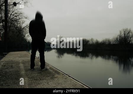 A mental health concept. A mysterious hooded figure, Looking across at a river on a bleak winters day, with a blurred edit. Stock Photo