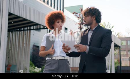 Indian Arabian male man businessman holding papers documents corporate discuss result plans with African American woman colleague businesswoman Stock Photo