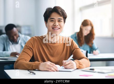 Asian male student sitting at desk writing in classroom Stock Photo