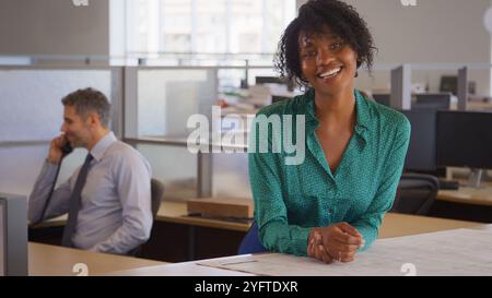 Portrait Of Smiling Female Architect In Modern Open Plan Office Studying Building Plans Or Drawings Stock Photo