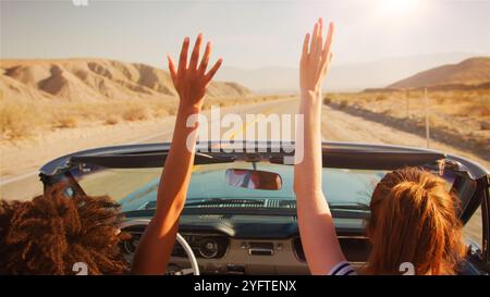 Two Female Friends Driving Classic Open Top Car On Road Trip In Countryside With Hands In The Air Stock Photo