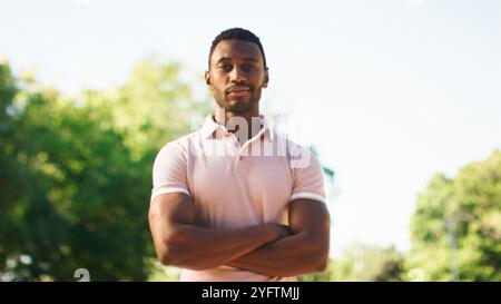 A young man stands outdoors with his arms crossed, exuding confidence. He is dressed casually in a light-colored shirt and is surrounded by lush green Stock Photo