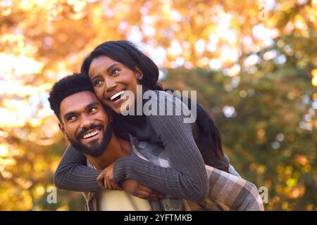 Loving Young Couple With Man Giving Woman Piggyback Against Background Of Autumn Leaves Stock Photo