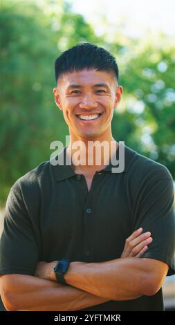 A young man stands confidently outdoors, smiling brightly with his arms crossed. The background features lush greenery, indicating a vibrant and warm Stock Photo