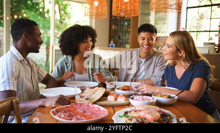 A group of four friends engages in cheerful conversation while sharing a variety of delicious dishes around a beautifully set dining table. The ambian Stock Photo