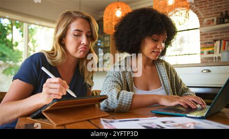 Two women collaborate at a kitchen table, one focused on writing with a tablet while the other types on a laptop. Natural light streams through large Stock Photo