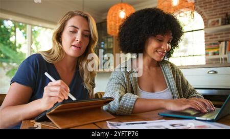 Two women are engaged in a productive collaboration at a wooden table. One woman is using a digital tablet while the other types on her laptop, both s Stock Photo