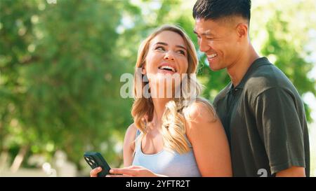 Couple shares a joyful moment while using a smartphone in a sunny park. Stock Photo