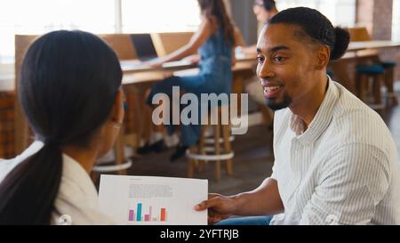 Two Young Businesspeople Meeting In Office Discussing Documents With Colleagues In Background Stock Photo