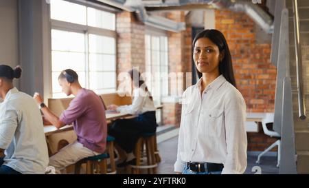 Portrait Of Young Smiling Businesswoman Working In Open Plan Office With Colleagues In Background Stock Photo
