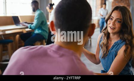 Two Young Businesspeople Meeting Having Discussion In Modern Office With Colleagues In Background Stock Photo