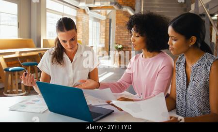 Three Young Businesswomen Meeting In Modern Office Sitting Around Table Working On Laptop Together Stock Photo