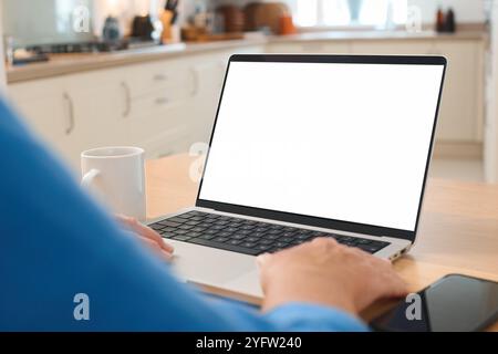 A person is seated at a kitchen table, focused on a laptop computer with a blank screen. The bright kitchen features warm wood cabinetry and natural l Stock Photo