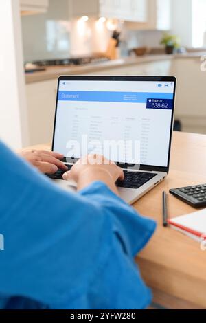 A person is working diligently on a laptop in a sunlit kitchen, analyzing their financial statement. The workspace features a calculator, a notepad, a Stock Photo
